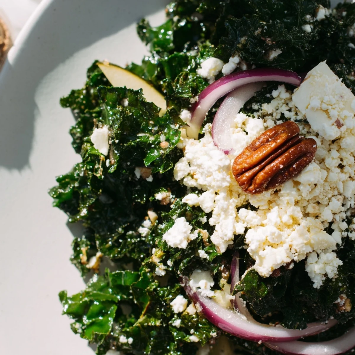 A large serving bowl of Winter Kale Salad with Pecans and Cranberries set on a rustic wooden table. The vibrant green kale is tossed with shiny toasted pecans, bright red cranberries, and thin apple slices, all lightly coated in zesty vinaigrette. A wooden spoon rests in the salad, ready to serve four people.