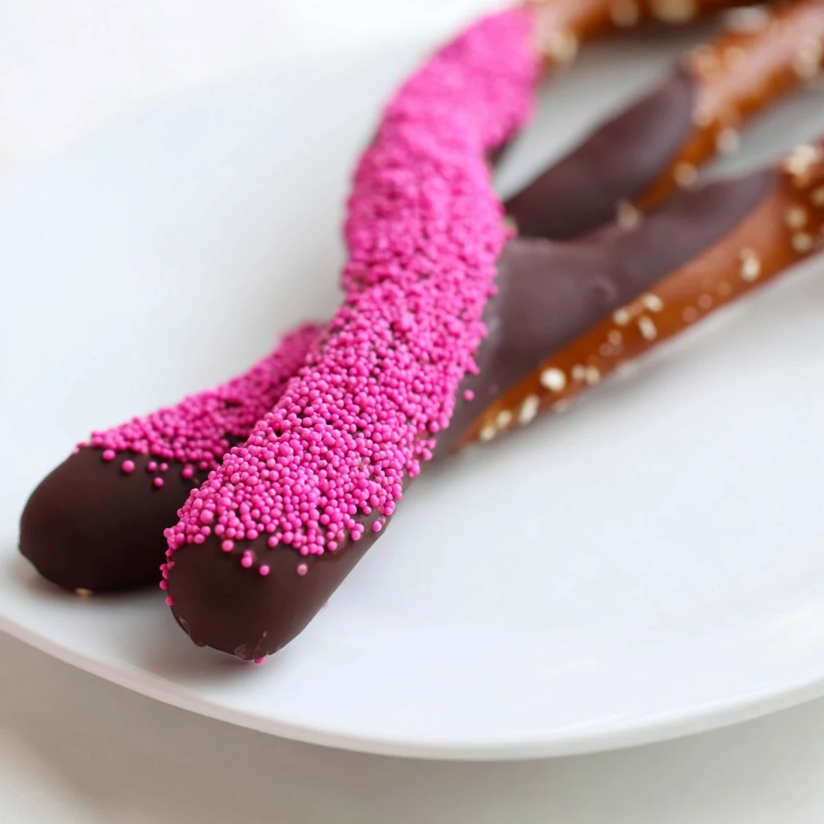 Salty pretzel rods dipped in rich chocolate and topped with pink sprinkles, set on parchment paper for a clean presentation.