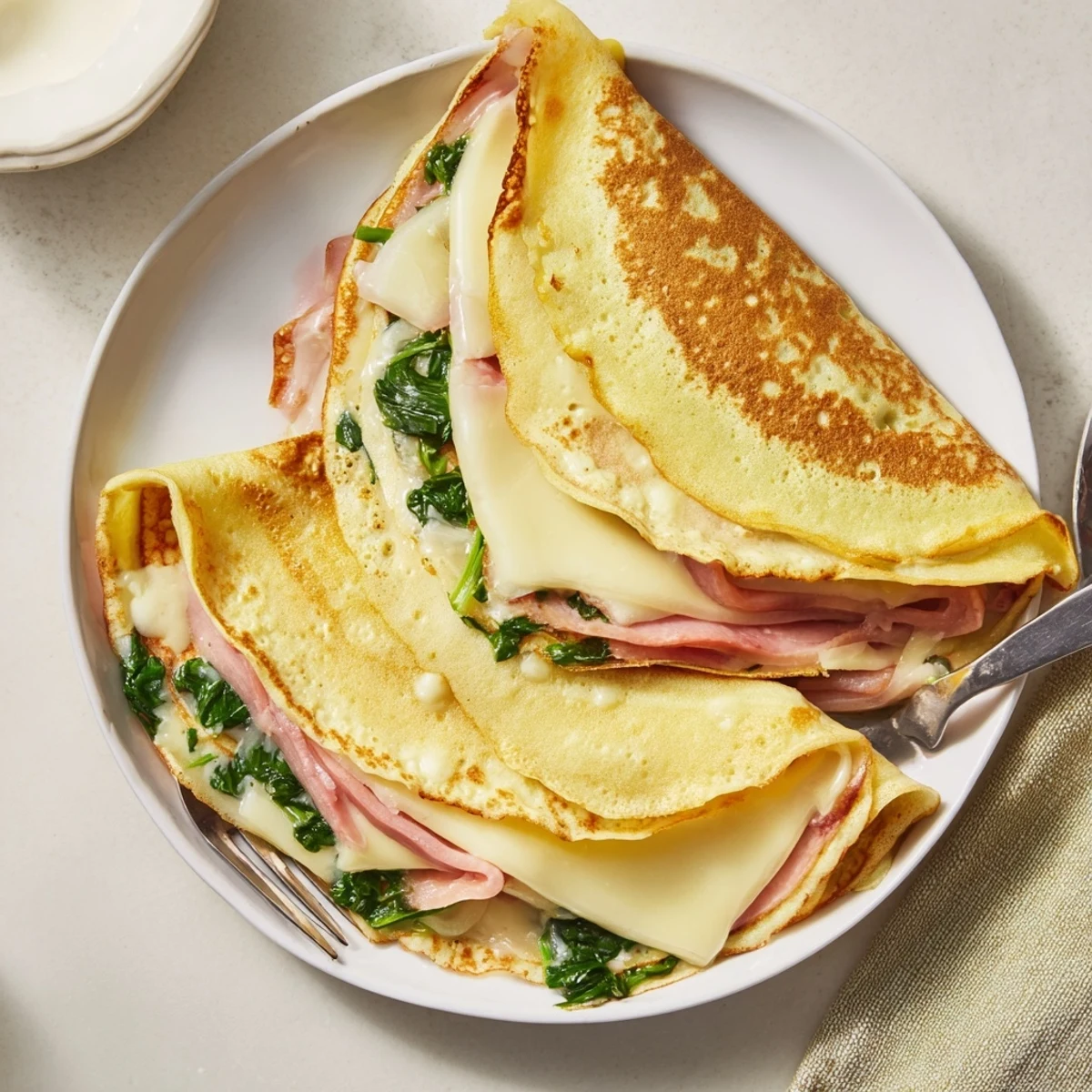 Golden savory crepes folded with melted Gruyère cheese and fresh spinach filling on a white plate